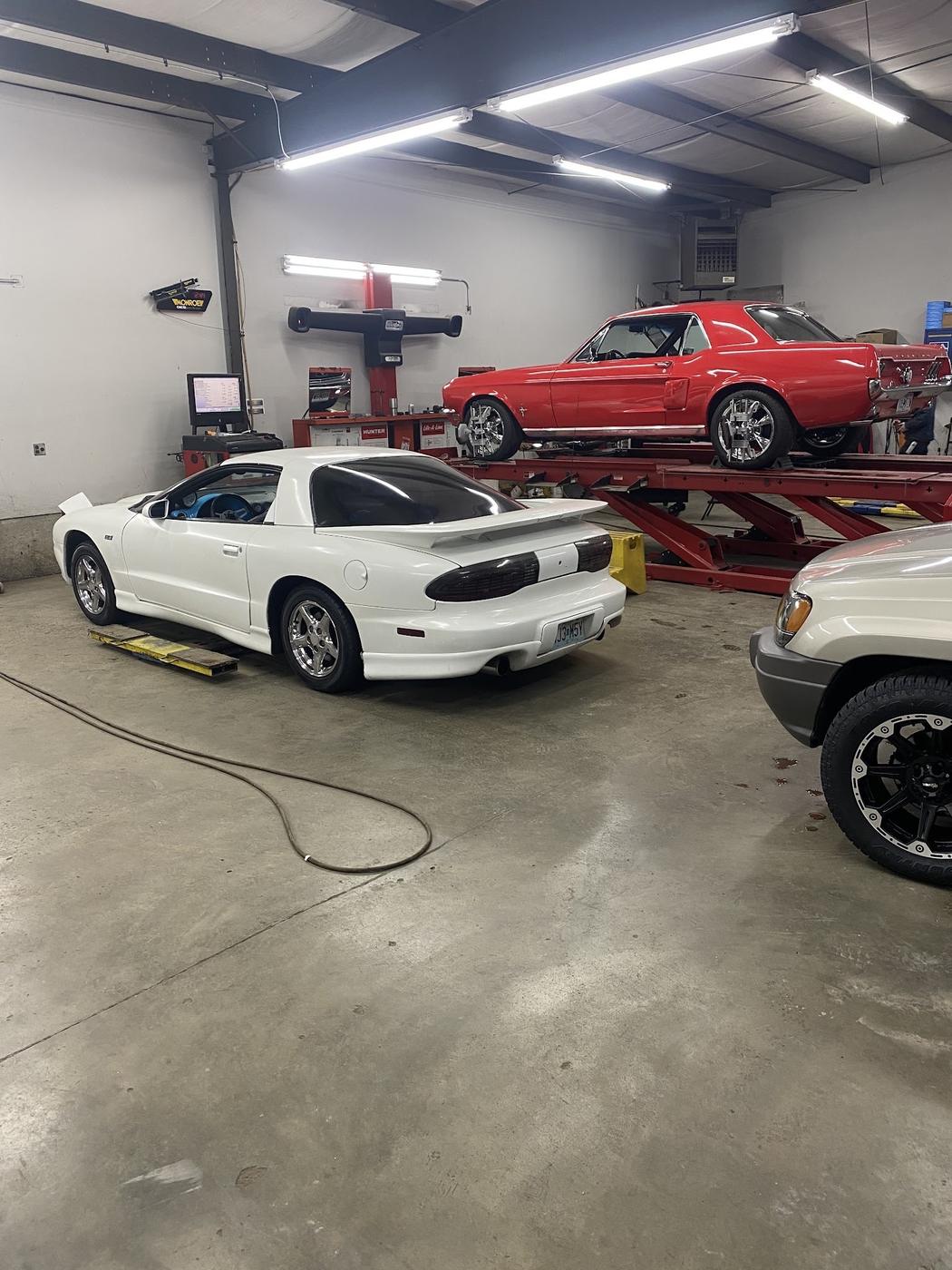 Inside the shop: a red Mustang on the alignment rack and a white Firebird on the floor