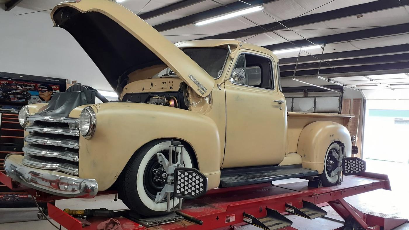 A classic pickup with its hood up on the alignment rack, targets clamped to the wheels